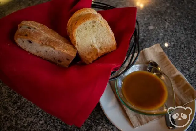 Bread wrapped in a red, cloth napkin in a basket with a bowl of tomato based sauce. 