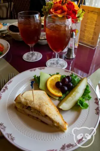 Two glasses of iced tea with a sandwich and a side of fruit on a plate.