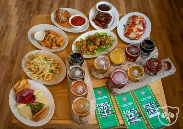 Table full of food with a charcuterie platter, fries, fried artichoke hearts, meatball sandwich, ribs, chicken thighs, pizza, and 3 flights of beers. 